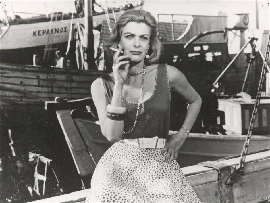 Melina Mercouri na dobové fotografie, v ruce drží cigaretu a za ní jsou lodě v přístavu